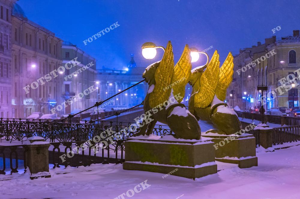 На фотографии изображен Банковский мост через канал Грибоедова в Санкт-Петербурге. Этот пешеходный мост известен своими скульптурами крылатых грифонов с фонарями. Зимний снег покрывает мост и фигуры, добавляя атмосферы уюта и сказочности. Фон подсвеченных зданий и голубоватое небо создают особенное вечернее настроение.